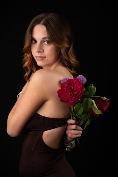 Aura in abito nero su fondale nero, con rose rosse dietro la schiena
